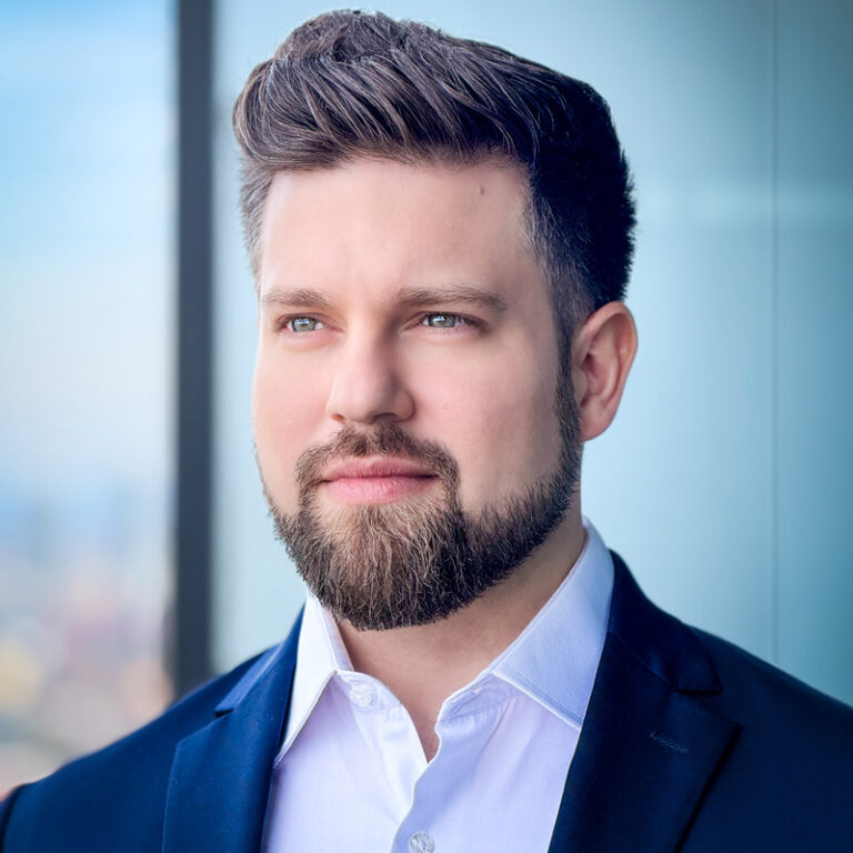 Andrés Sevilla wearing a white collared shirt inside a navy blue blazer and looking up and off to the left. Andrés has short brown hair and a beard and mustache.