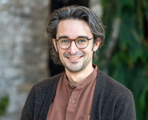 Headshot of Ben Lee Taylor with glasses, a brown collared shirt inside a dark grey cardigan, and a smile on his face.