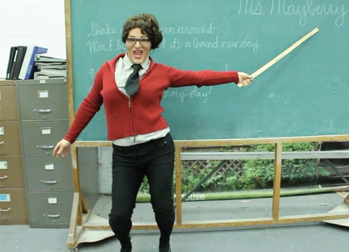 Dr. Tommy in drag in front of a chalkboard with a metre stick in their hand pointing at the name "Ms. Mayberry" in cursive writing. Tommy is wearing "Sarah Palin"-esque drag.