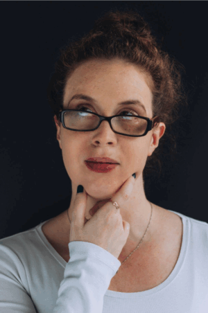 Headshot of Dr. Lisa Endersby in a long-sleeved, white top holding her chin inquisitively with her right hand and looking off to the top left. Lisa is wearing black framed glasses and has her hair in a bun on the top of her head.