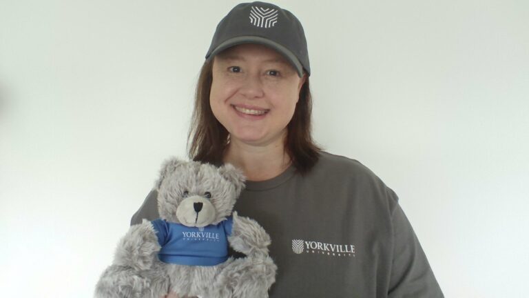 Dr. Louise Olivier wearing a grey Yorkville U sweater, a grey ball cap with the Yorkville U logo on it, and a holding a grey teddy bear who is wearing a blue t-shirt with the Yorkville U logo on it.