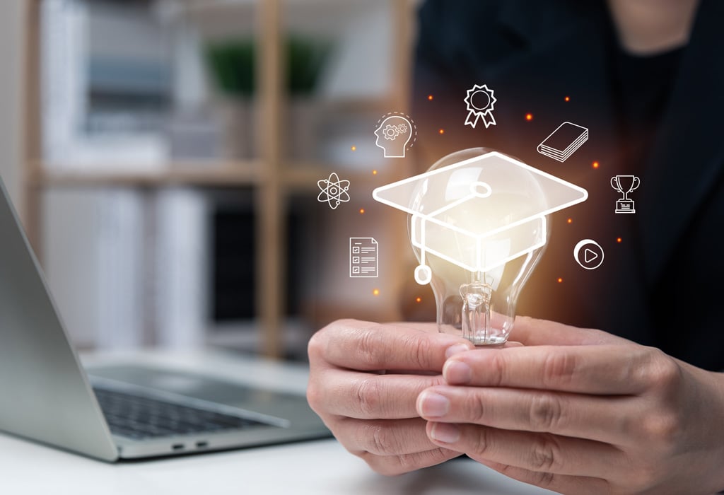 Hands in front of a laptop holding a lightbulb with a digital graduation cap on it and teaching and learning icons surrounding it.