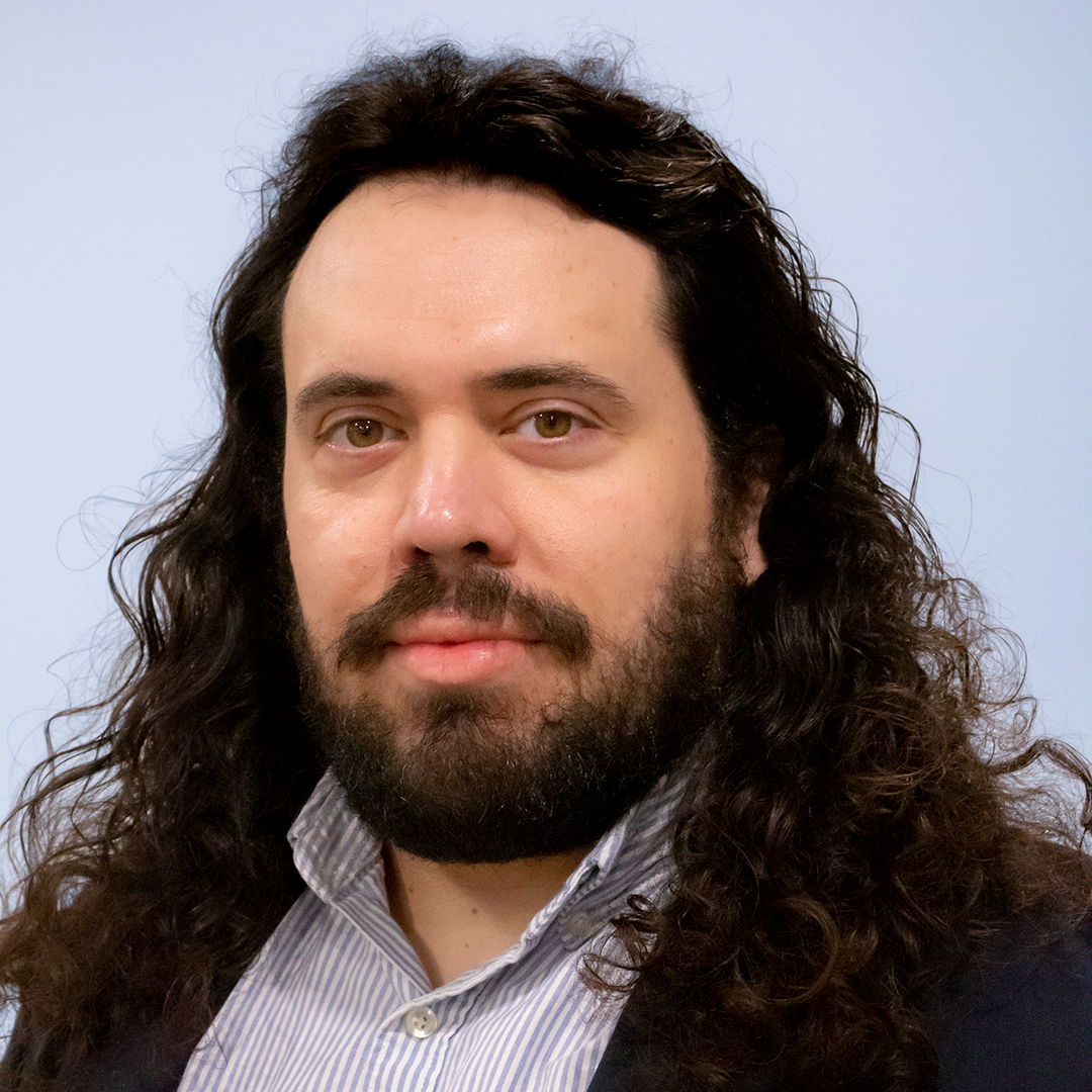 Professional headshot of Bruno Lyra, who is wearing a collared shirt under a blazer and has long, wavy dark hair and a dark beard and mustache.