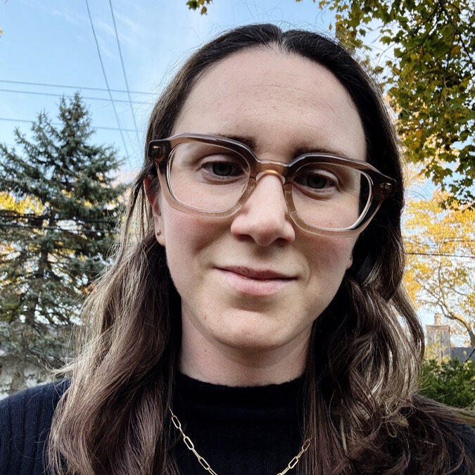 Sarah in front of pines trees and with long, shoulder-length light brown hair and glasses.